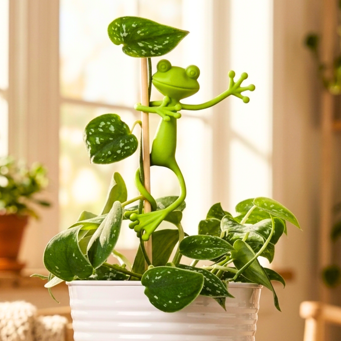Tuteur décoratif Mamie-Frog en forme de grenouille verte accrochée à une tige de plante en pot avec des feuilles de couleur rose